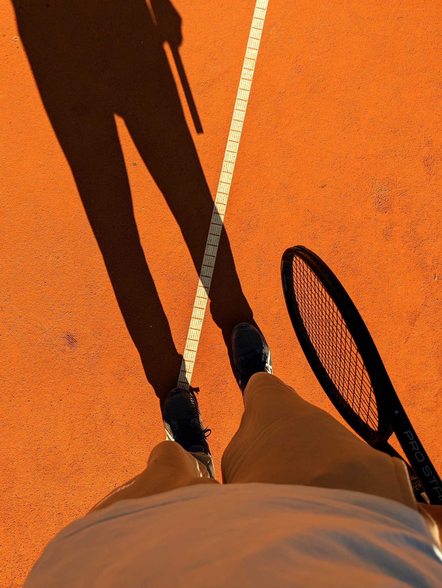 First-person view on a clay tennis court, racket and shadow on the orange surface