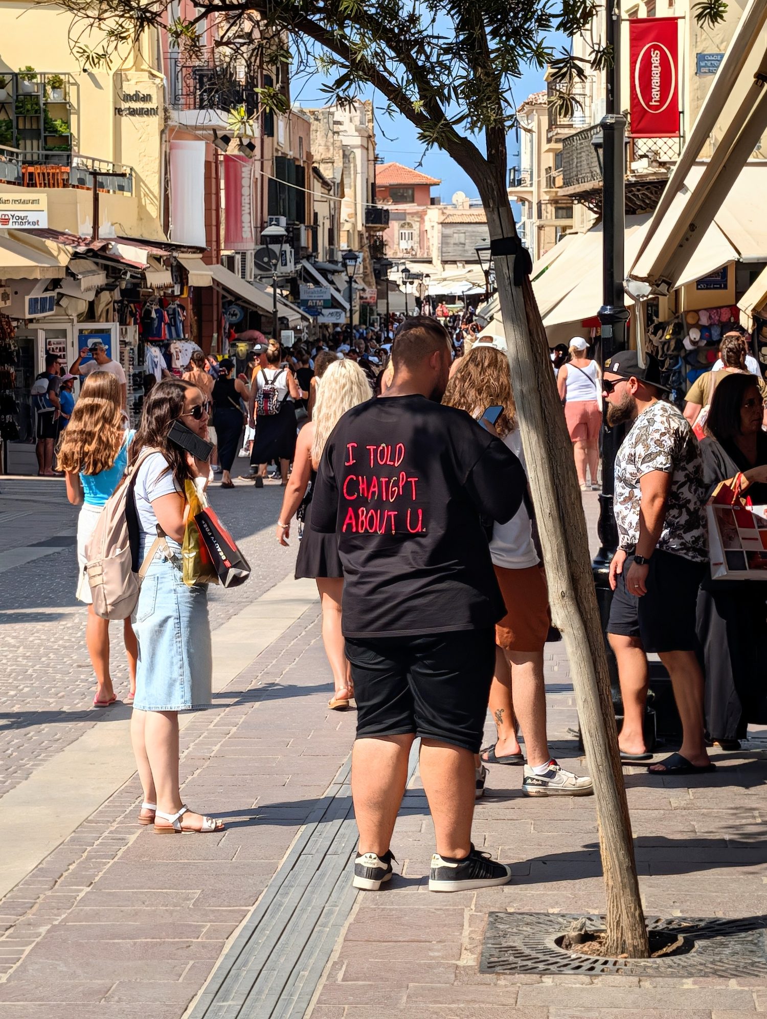 A busy pedestrian shopping street lined with shops and awnings. In the middle, a man walks away from the camera in a black T-shirt with pink handwritten lettering on the back reading "I TOLD CHATGPT ABOUT U."