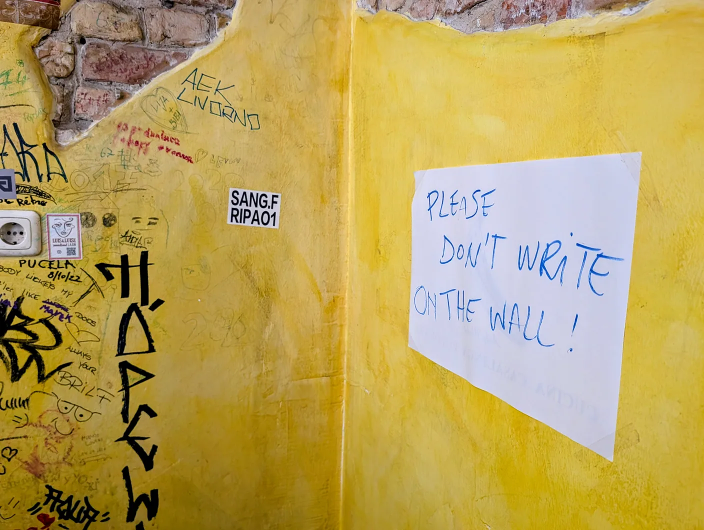 A yellow wall covered in graffiti beside a handwritten paper sign saying Please don't write on the wall!