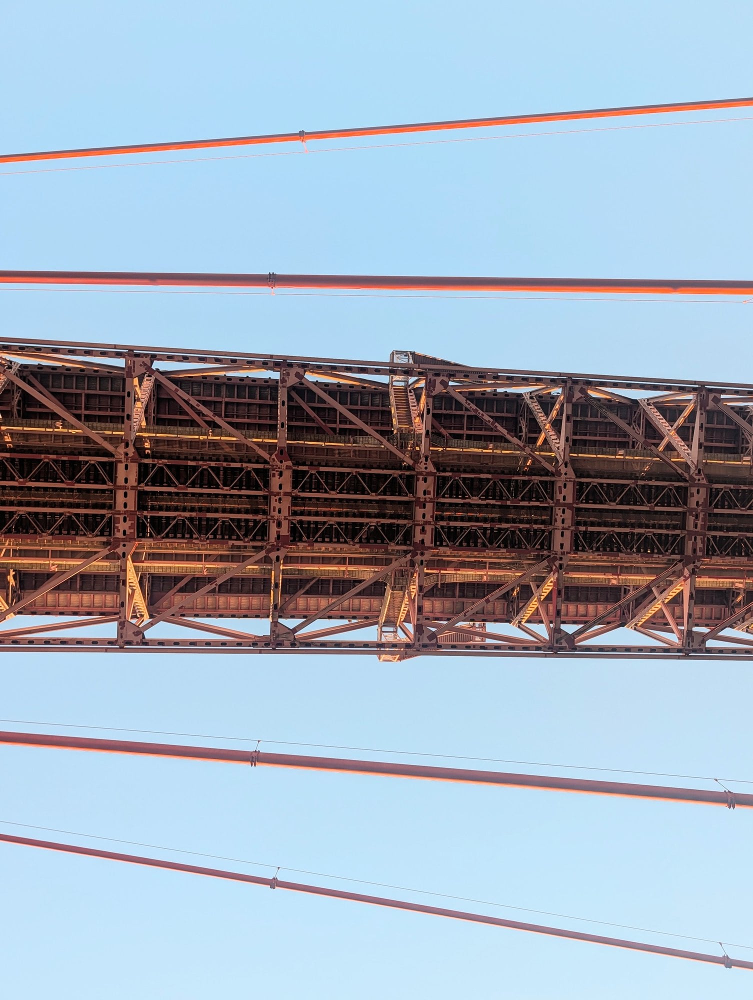 The underside of the Ponte 25 de Abril in Lisbon seen from below at golden hour: red steel truss deck with its lattice of girders and stairways, suspension cables crossing a pale blue sky