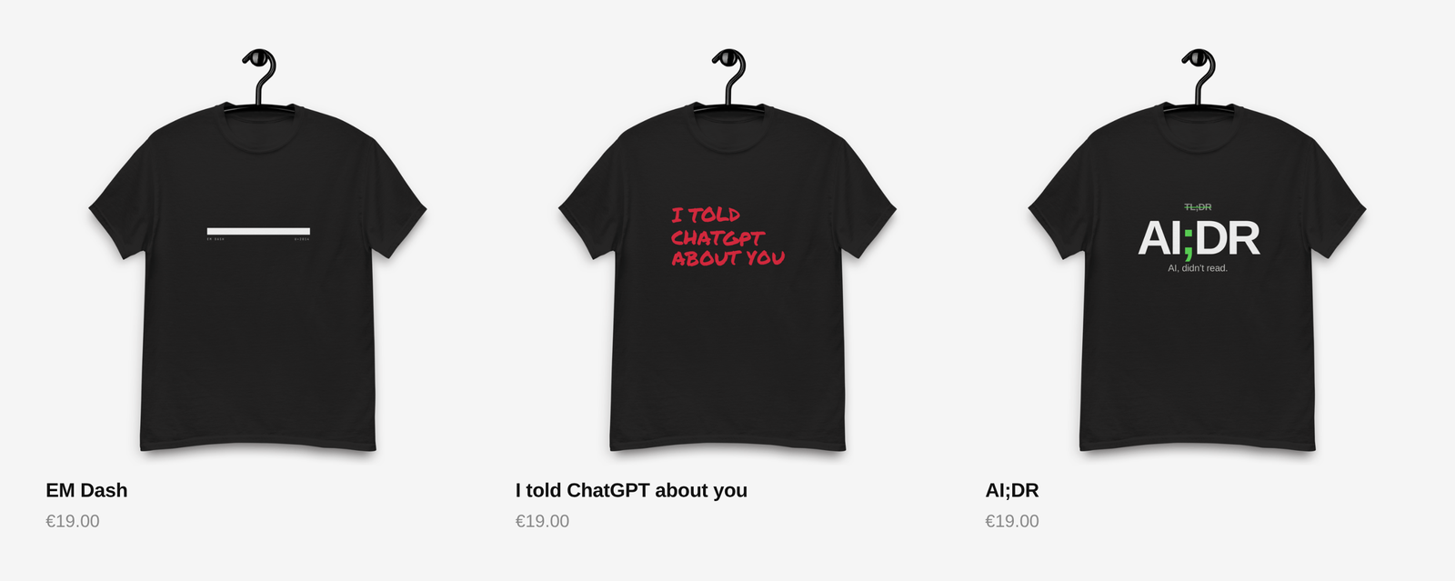 Three black T-shirts on hangers against a light grey background: EM Dash with a white bar, I told ChatGPT about you in red handwritten lettering, and AI;DR with a green semicolon. Each priced at 19 euro.