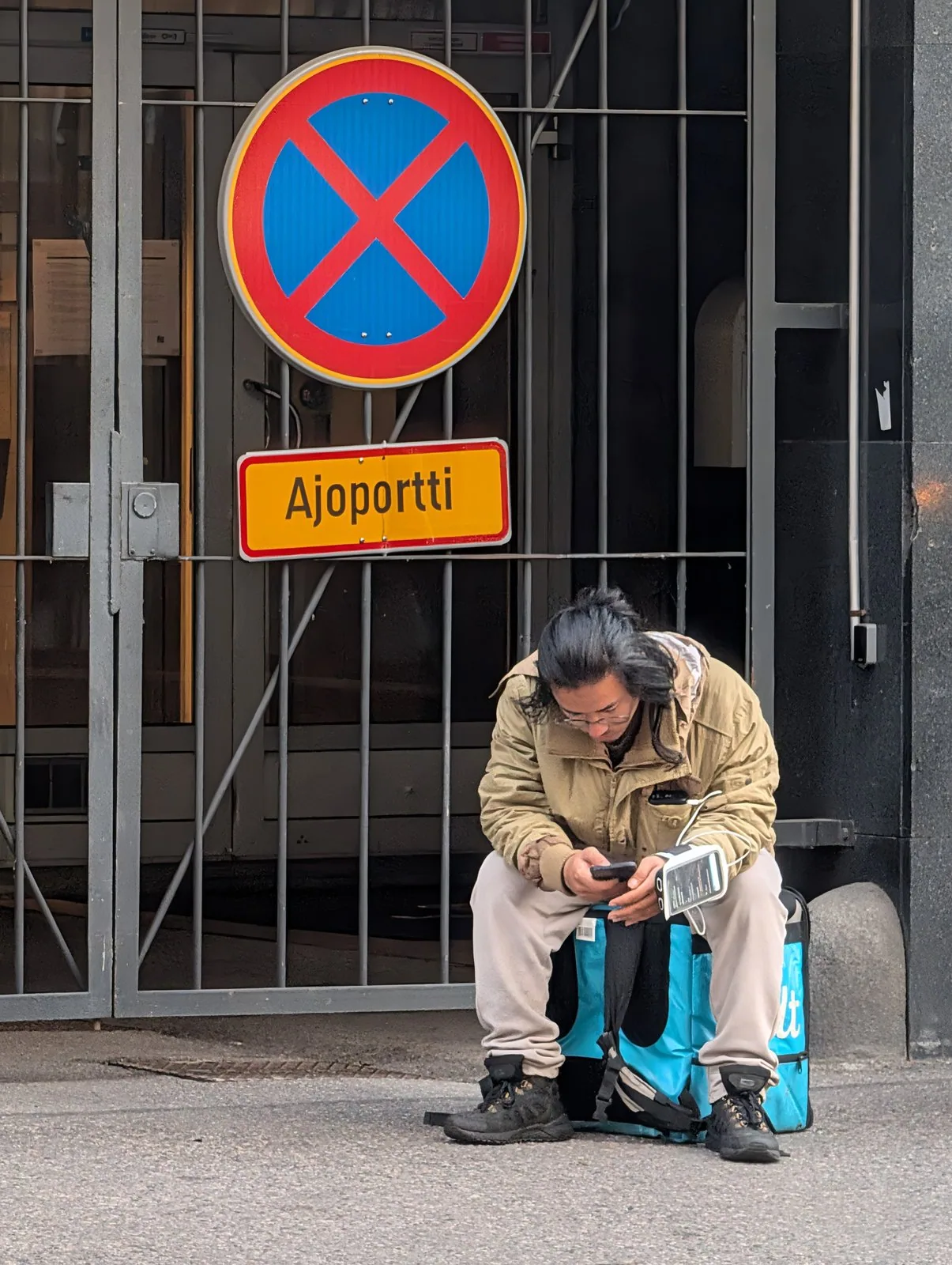 A Wolt courier sitting on his delivery bag checking his phone, beneath a no-stopping sign marked “Ajoportti”